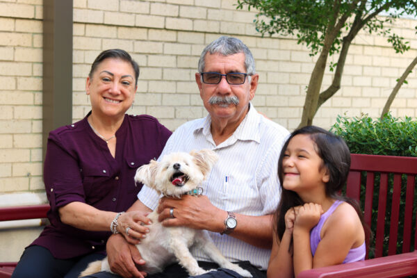 A family sits on a bench outdoors with their small, fluffy dog. The dog is held by the person in the center, who is wearing glasses and a striped shirt. A person in a purple shirt is to the left, and a child in a purple top is to the right.