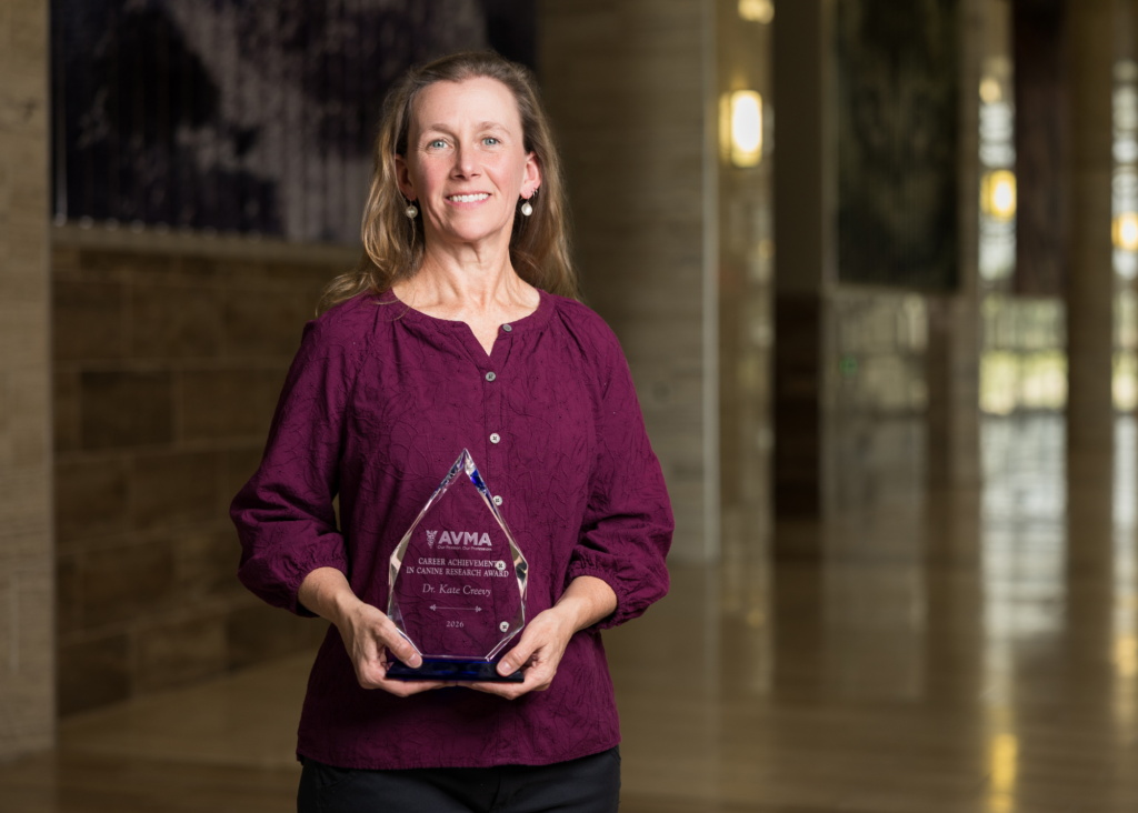 A woman named Kate Creevy, wearing a maroon blouse, holds a glass award that reads "AVMA Career Achievement in Canine Research Award - Dr. Kate Creevy 2026."